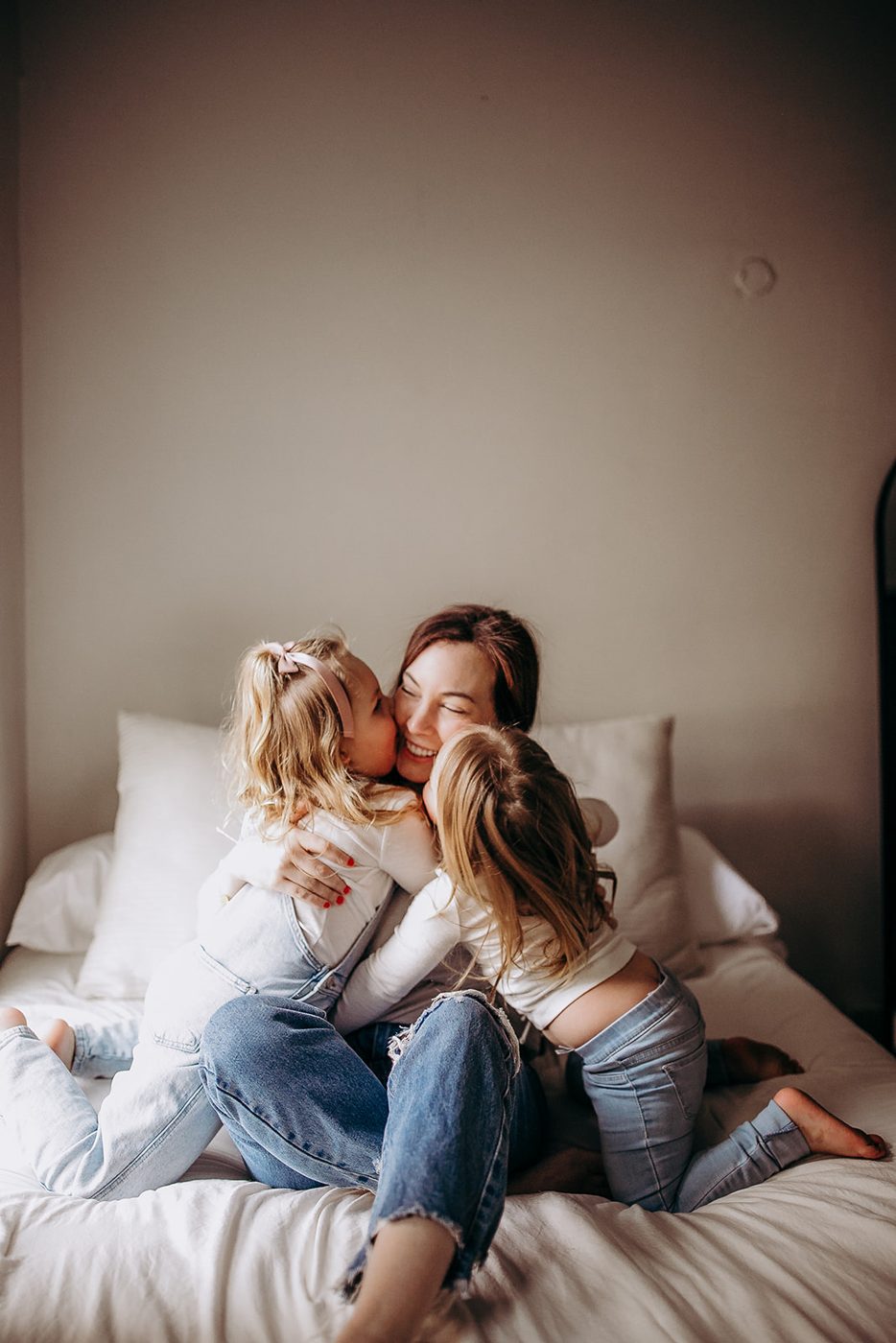 Harley laughing on a white bed while her two young daughters kiss her on both cheeks